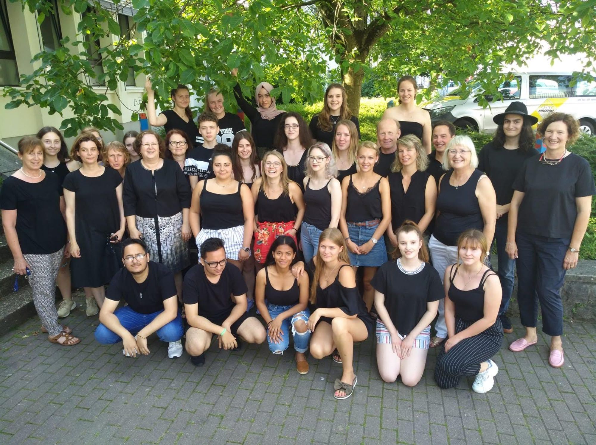 Auf dem Bild sieht man eine Menschengruppe die in vier Reihen auf dem Pausenhof aufgeteilt sind.
